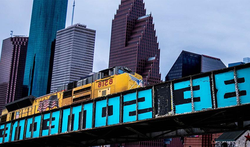 Picture of a train passing over the top of "Be Someone" painting with Houston buildings in the background.